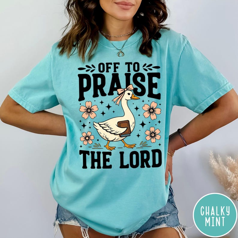 Puede incluir: Camiseta azul claro con el texto "OFF TO PRAISE THE LORD". Presenta un dise&ntilde;o de un ganso con lazo y libro, rodeado de flores rosas. La camiseta es de corte holgado, adecuada para uso casual.