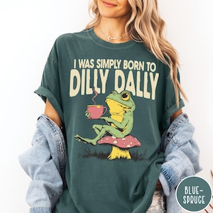 May include: Teal-colored t-shirt with the text "I WAS SIMPLY BORN TO DILLY DALLY." A cartoon frog is seated on a mushroom, holding a pink cup. The frog is green and yellow. The shirt has rolled-up sleeves.