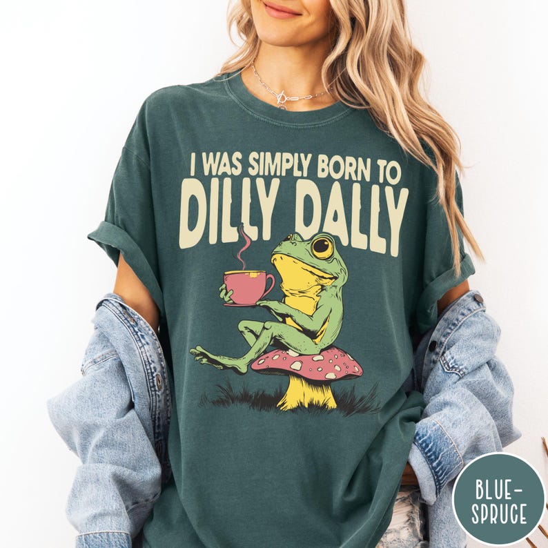 May include: Teal-colored t-shirt with the text "I WAS SIMPLY BORN TO DILLY DALLY." A cartoon frog is seated on a mushroom, holding a pink cup. The frog is green and yellow. The shirt has rolled-up sleeves.
