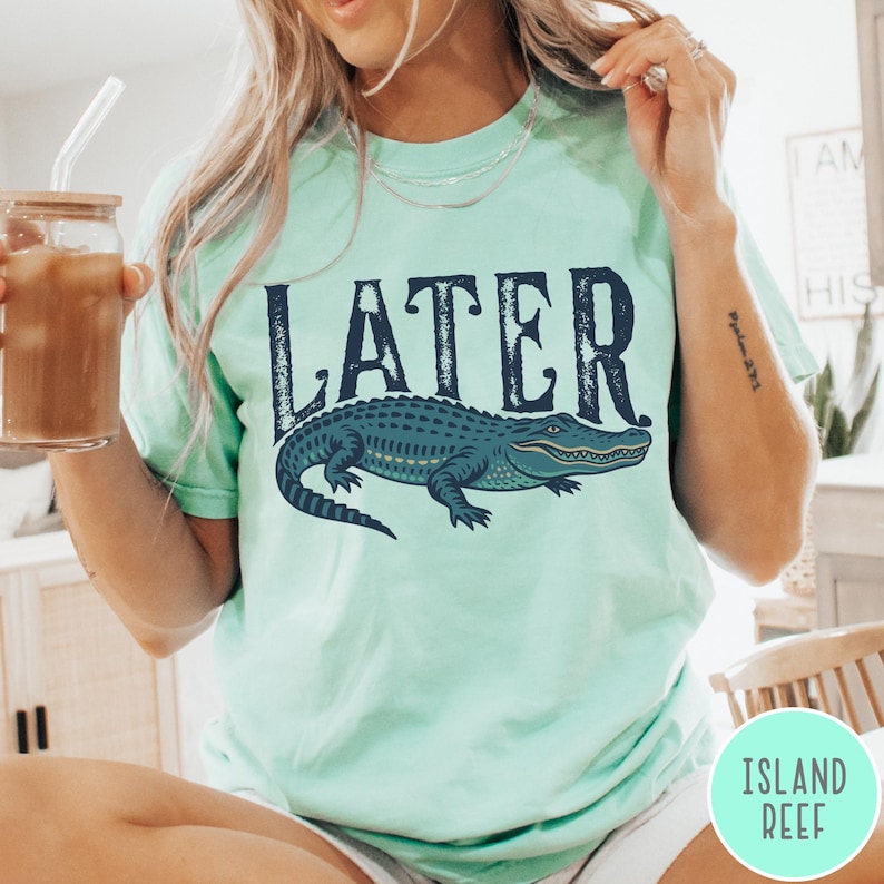 May include: A light green t-shirt with the word "LATER" in dark blue above a detailed illustration of a blue alligator. The shirt has a relaxed fit and short sleeves. The text "ISLAND REEF" is in a circle on the bottom right.