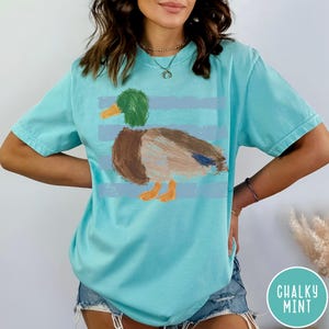 May include: A light turquoise short-sleeve t-shirt featuring a central graphic of a mallard duck rendered in a textured, painterly style. The duck has a green head, yellow beak, brown body, and orange feet, set against horizontal light blue stripes.
