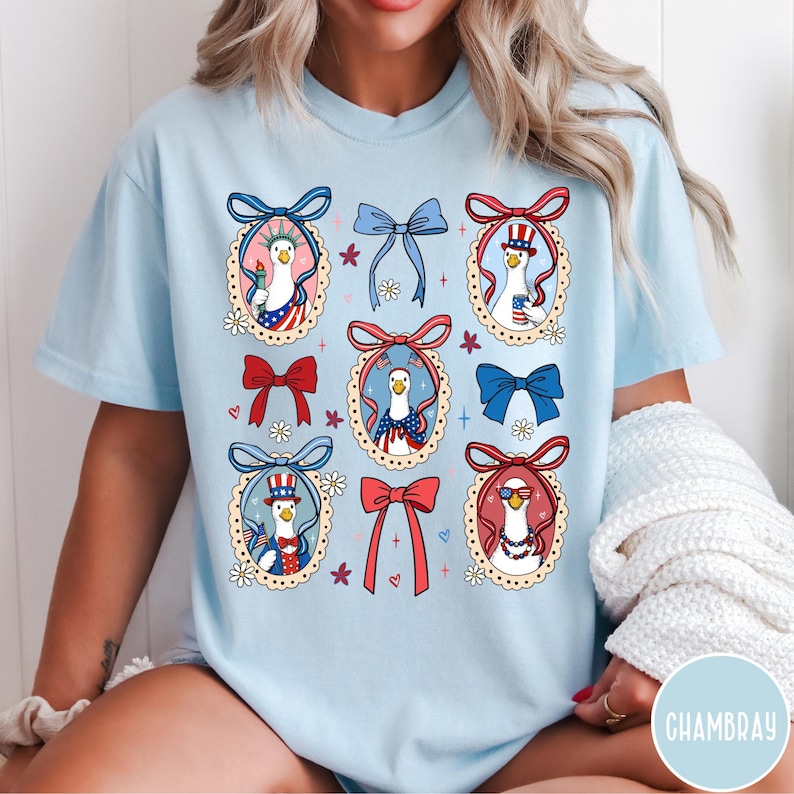 May include: Light blue t-shirt featuring a patriotic design with cartoon geese wearing hats and sunglasses, surrounded by red, white, and blue bows and floral accents. The design is repeated across the shirt.