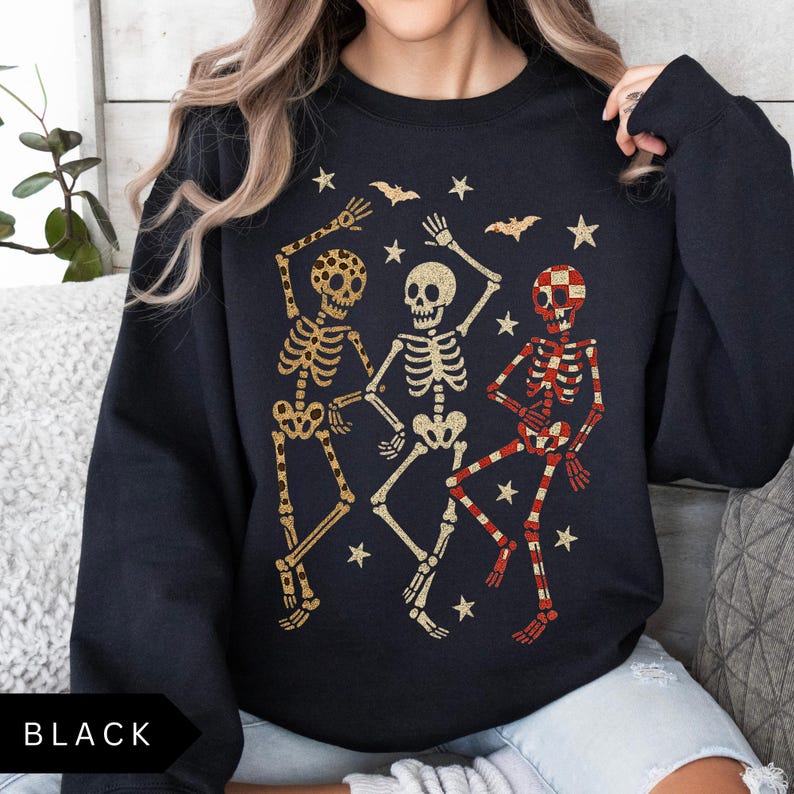 Puede incluir: Sudadera negra con tres esqueletos bailando en patrones dorados y a cuadros rojos, con estrellas y murci&eacute;lagos. La palabra "BLACK" est&aacute; impresa en la parte inferior. Un dise&ntilde;o festivo para Halloween.
