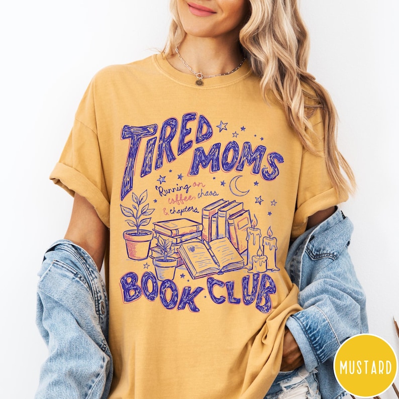 May include: Mustard-colored t-shirt with the text "Tired Moms Book Club" in purple. The design includes illustrations of books, candles, plants, and stars. The shirt also has the text "Running on coffee, chaos & chapters."