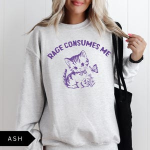 以下が含まれることがあります： 紫色の文字で「Rage Consumes Me」とプリントされたヘザーグレーのスウェットシャツ。テキストの下には、蝶が飛んでいる上に、リボンをつけた漫画の猫が描かれています。