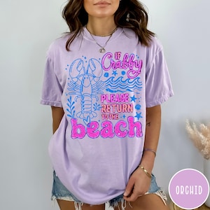 Puede incluir: Una camiseta lila con el texto "If Crabby Please Return to the Beach" en rosa y azul. El dise&ntilde;o incluye una langosta y olas. Esta camiseta informal es ideal para unas vacaciones en la playa.