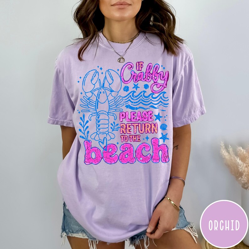 Puede incluir: Una camiseta lila con el texto "If Crabby Please Return to the Beach" en rosa y azul. El dise&ntilde;o incluye una langosta y olas. Esta camiseta informal es ideal para unas vacaciones en la playa.