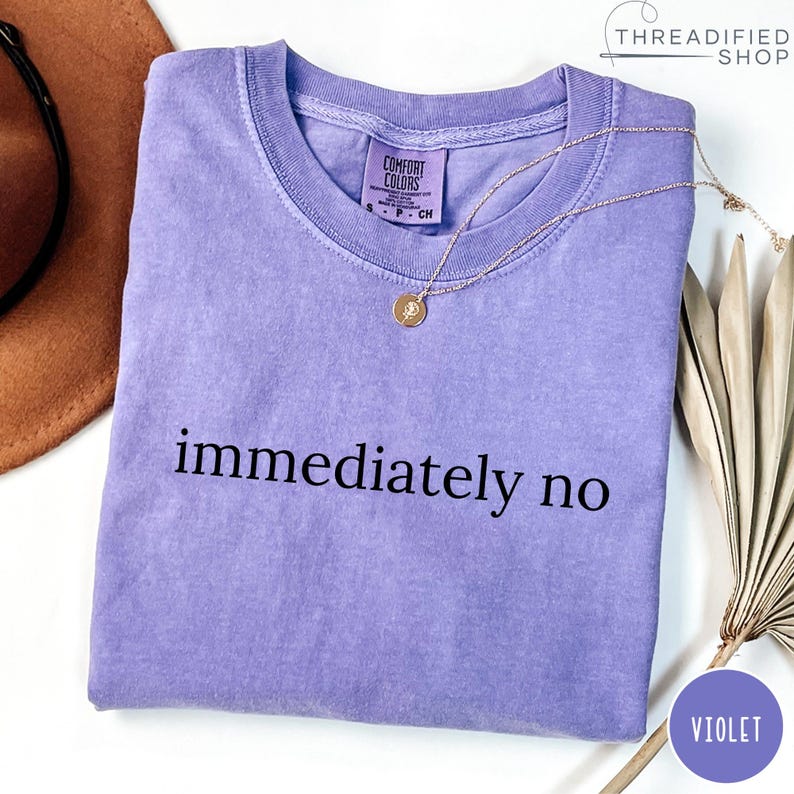 May include: A violet-colored t-shirt with the words "immediately no" printed in black. A gold necklace with a pendant rests on the shirt. The shirt is from the Threadified Shop.