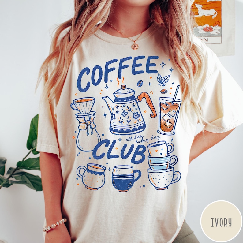 May include: Ivory-colored t-shirt with a blue and orange graphic that reads "Coffee Club." The design includes coffee-related illustrations such as a teapot, coffee cups, and a glass of iced coffee. The text "all day, every day" is also included.