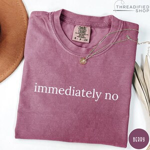 May include: Berry-colored t-shirt with the words "immediately no" printed in white. The shirt has a crew neck and short sleeves. A gold necklace with a pendant is draped across the shirt. The shirt is from the Comfort Colors brand.