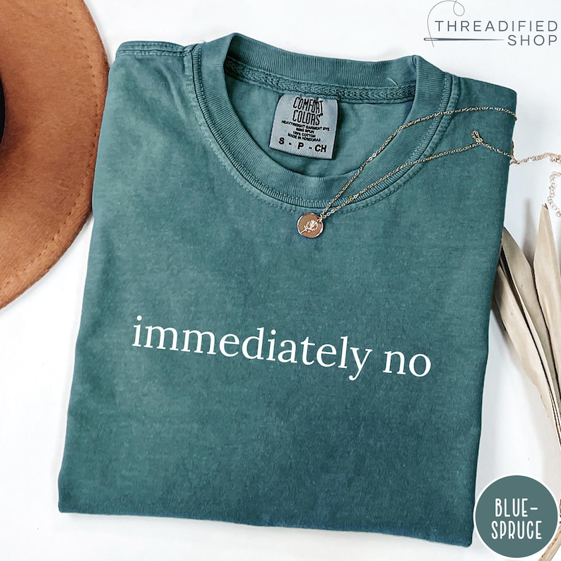May include: A teal Comfort Colors t-shirt with the words "immediately no" in white. A gold necklace with a pendant rests on the shirt. The shirt is folded and placed on a white surface.