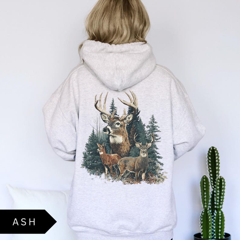 Op de afbeelding: Lichtgrijze hoodie met een hertenafbeelding op de rug. De afbeelding toont een bok met grote geweien, twee herten en groenblijvende bomen. Het woord "ASH" is gedrukt op een zwart, pijlvormig label.
