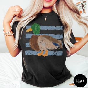 May include: A black short-sleeve t-shirt featuring a graphic of a mallard duck with a green head, brown body, and orange feet. The duck is layered over a background of horizontal, distressed light blue stripes. A small black circle with the text BLACK.