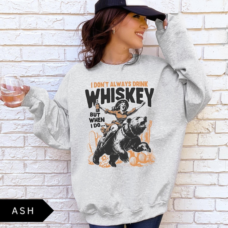 May include: A gray sweatshirt with the text 'I don't always drink whiskey but when I do...' and a cartoon image of a woman riding a bear. The woman is wearing a cowboy hat and holding a bottle of whiskey.
