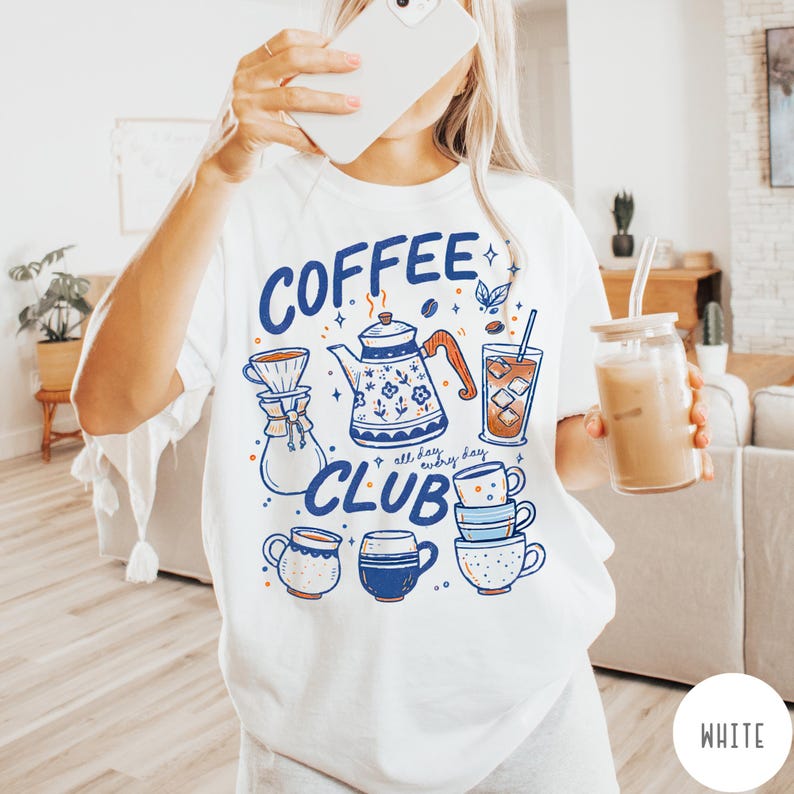 May include: White t-shirt with a blue and orange graphic that says "Coffee Club." The graphic includes a coffee pot, coffee cups, and a glass of iced coffee. The shirt is a casual, everyday wear item.