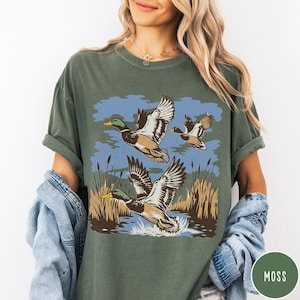 May include: Moss green t-shirt with a graphic of three ducks in flight over water and reeds. The ducks have brown, white, and green feathers with yellow beaks. The background includes blue sky and water. The shirt has a relaxed fit.