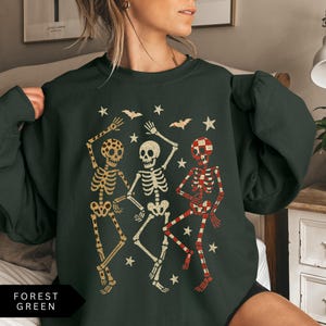 Puede incluir: Sudadera verde bosque con tres esqueletos bailando en beige y rojo, con estrellas y murci&eacute;lagos. Los esqueletos tienen diferentes patrones. El texto "FOREST GREEN" est&aacute; en la parte inferior.