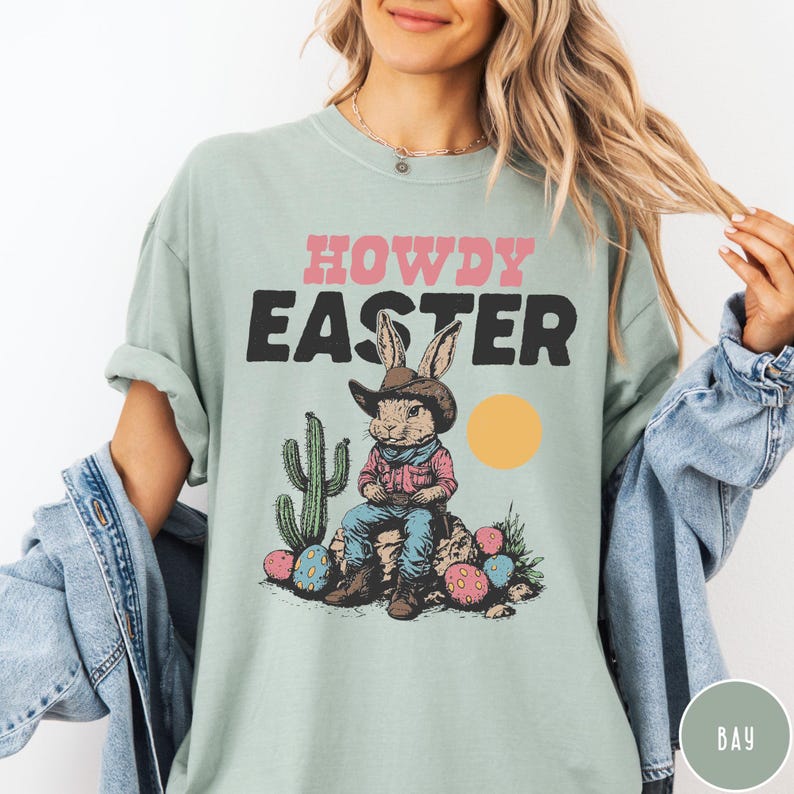 Puede incluir: Camiseta verde claro con las palabras "HOWDY EASTER" en rosa y negro. El gr&aacute;fico presenta un conejo con sombrero de vaquero, sentado en una roca con huevos de Pascua y un cactus. Tambi&eacute;n hay un c&iacute;rculo amarillo.