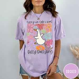 May include: A purple t-shirt with a white duck and colorful flowers. The text on the shirt reads "Sorry I'm late I was dilly dallying".