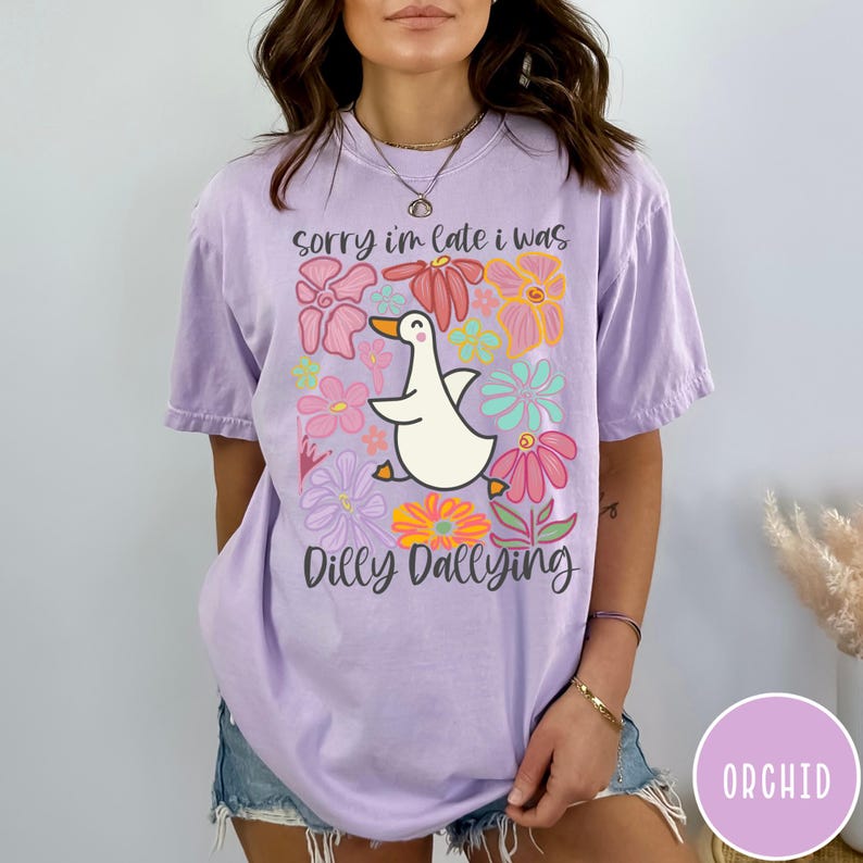 May include: A purple t-shirt with a white duck and colorful flowers. The text on the shirt reads "Sorry I'm late I was dilly dallying".