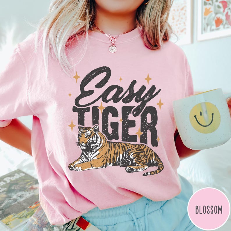 May include: Pink t-shirt with the text "Easy Tiger" in black lettering and a cartoon tiger illustration. The tiger is brown and orange with black stripes.