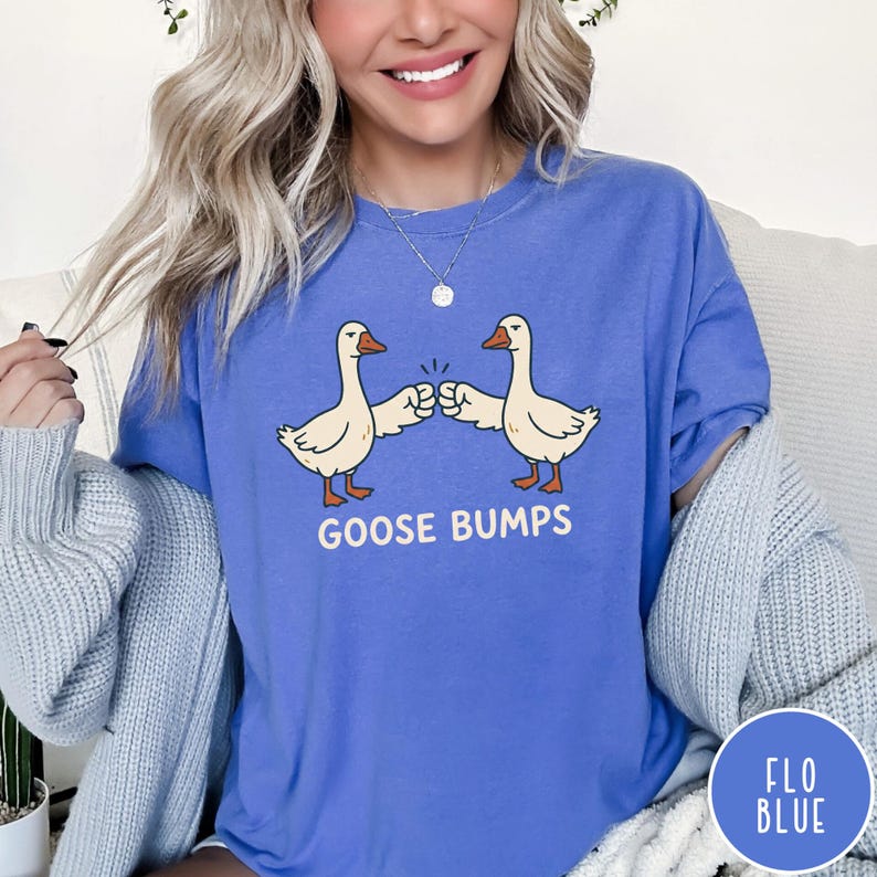 Puede incluir: Una camiseta de manga corta en color azul flo con un gr&aacute;fico de dos gansos blancos chocando los pu&ntilde;os. Debajo de los gansos, el texto GOOSE BUMPS est&aacute; impreso en letras may&uacute;sculas blancas. La camiseta tiene cuello redondo y corte holgado.