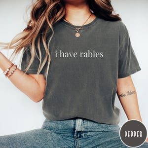 May include: A dark gray t-shirt with the text "i have rabies" in white. The shirt is a crew neck and appears to be made of a soft material. The model is wearing blue jeans and a bracelet.