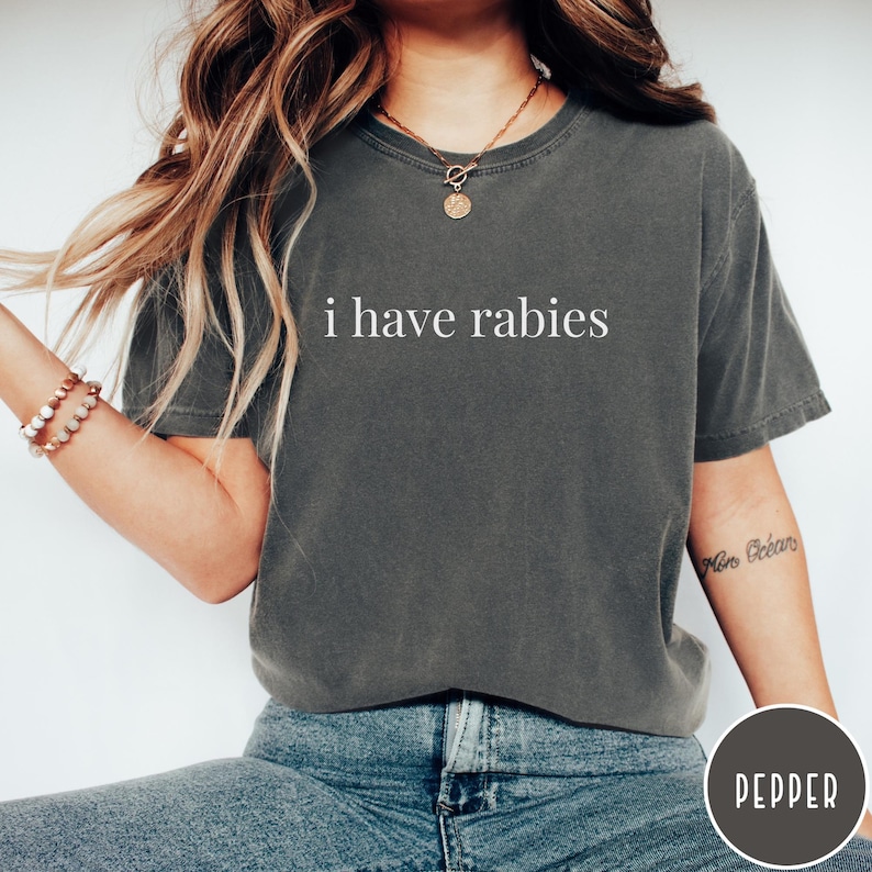 May include: A dark gray t-shirt with the text "i have rabies" in white. The shirt is a crew neck and appears to be made of a soft material. The model is wearing blue jeans and a bracelet.