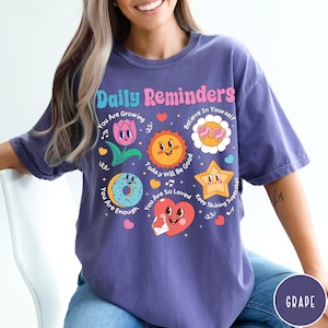 May include: A purple t-shirt with the words "Daily Reminders" in a colorful font. The shirt features various cartoon graphics, including a flower, sun, donut, star, and heart, each with positive messages like "You Are Enough" and "Believe In Yourself."