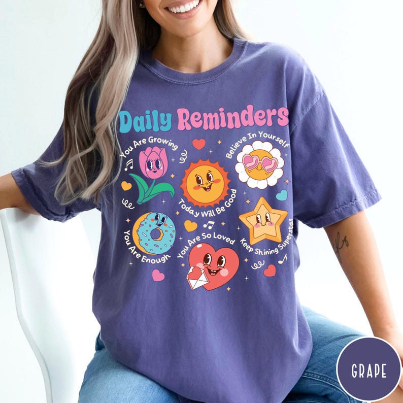 May include: A purple t-shirt with the words "Daily Reminders" in a colorful font. The shirt features various cartoon graphics, including a flower, sun, donut, star, and heart, each with positive messages like "You Are Enough" and "Believe In Yourself."