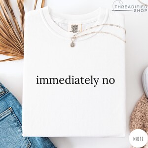 May include: White Comfort Colors t-shirt with the phrase "immediately no" in black. A silver necklace with a small pendant rests on the shirt. The shirt is folded, with a tag visible.