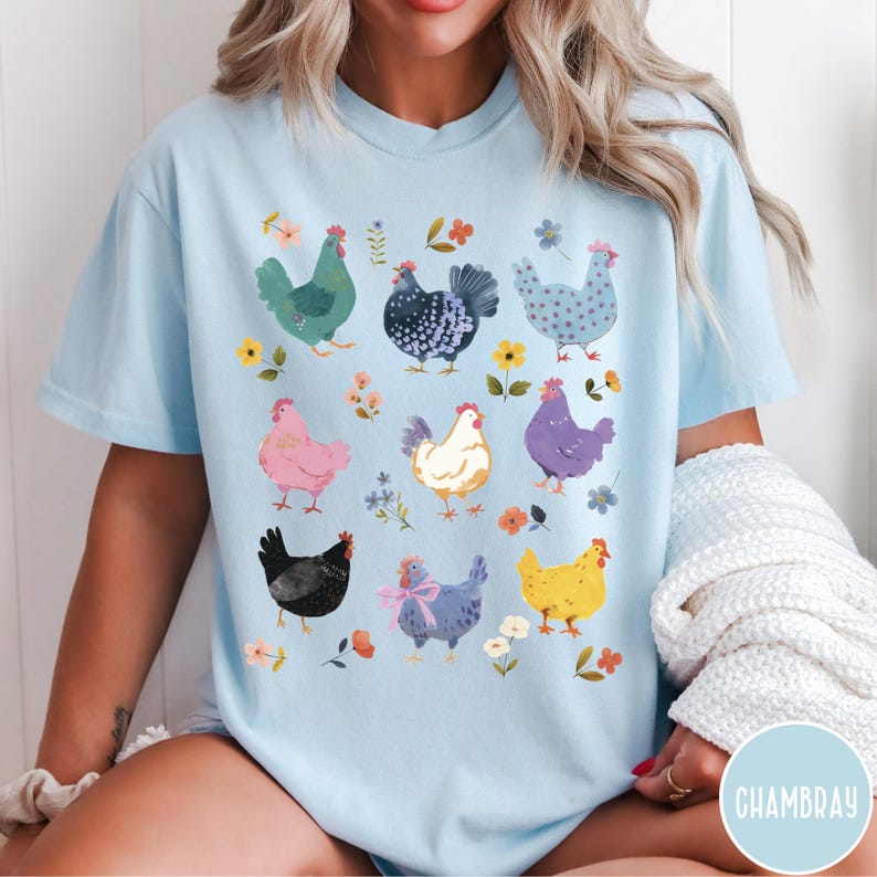 K&ouml;nnte beinhalten: Ein hellblaues T-Shirt mit einem bunten Grafikdruck von H&uuml;hnern und Blumen. Das Shirt hat einen Rundhalsausschnitt und kurze &Auml;rmel.