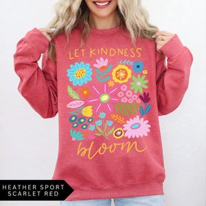 May include: A red heather sport sweatshirt with the text "Let Kindness Bloom" in white and yellow lettering. The text is surrounded by a colorful floral design.