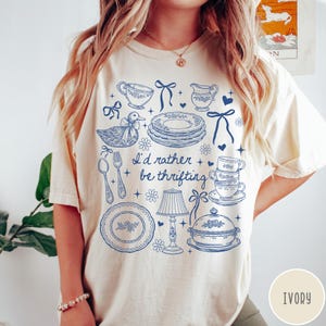 May include: Ivory t-shirt featuring a blue illustration of kitchenware, such as plates, cups, and a duck. The text "I'd rather be thrifting" is printed on the shirt. The t-shirt is a casual, comfortable style.