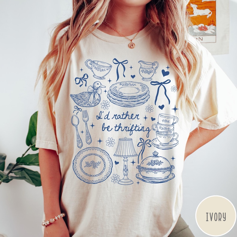 May include: Ivory t-shirt featuring a blue illustration of kitchenware, such as plates, cups, and a duck. The text "I'd rather be thrifting" is printed on the shirt. The t-shirt is a casual, comfortable style.