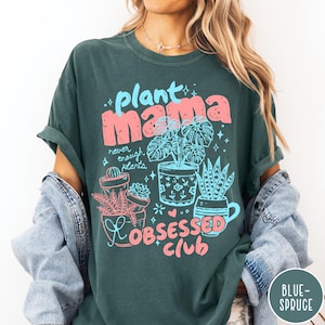 Puede incluir: Camiseta verde oscuro con las palabras "plant mama" en rosa y azul, junto con ilustraciones de plantas en macetas. Tambi&eacute;n presenta el texto "never enough plants" y "obsessed club". La camiseta tiene mangas enrolladas.