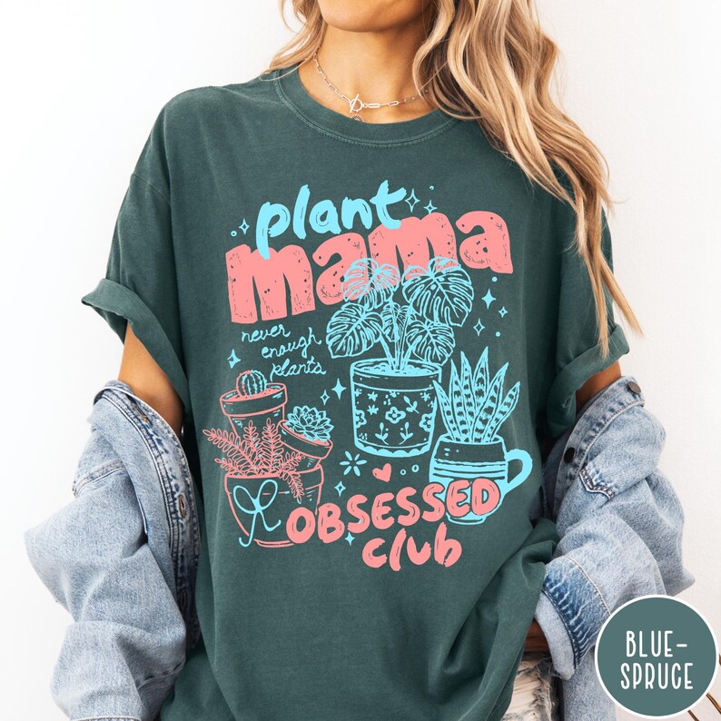 Puede incluir: Camiseta verde oscuro con las palabras "plant mama" en rosa y azul, junto con ilustraciones de plantas en macetas. Tambi&eacute;n presenta el texto "never enough plants" y "obsessed club". La camiseta tiene mangas enrolladas.