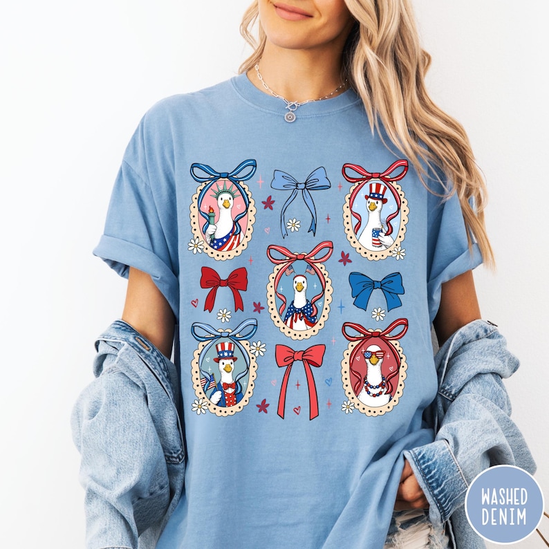 May include: Light blue t-shirt featuring a patriotic design with cartoon geese in red, white, and blue bow-framed portraits. The geese are wearing hats and sunglasses, and holding flags. The shirt has a relaxed fit and short sleeves.