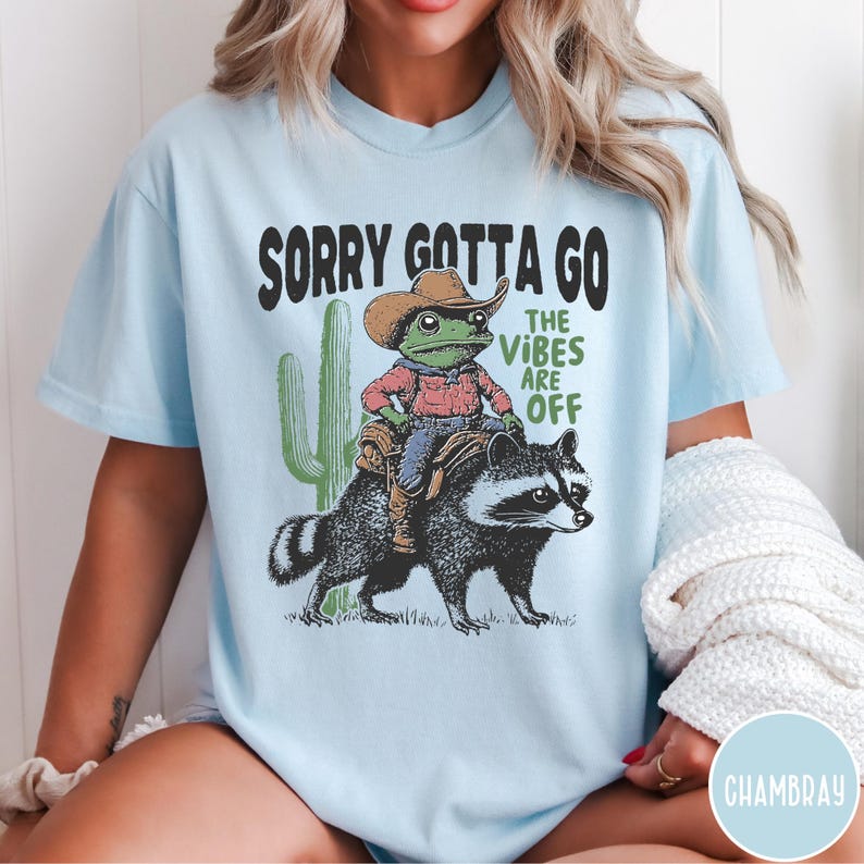 May include: Light blue t-shirt with a graphic featuring a frog in a cowboy hat riding a raccoon, with a cactus and the text "SORRY GOTTA GO THE VIBES ARE OFF". The design is printed on a light blue tee.