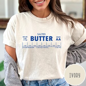 May include: A white t-shirt with a blue graphic of a stick of butter with the text "SALTED BUTTER GRADE AA" and a measurement guide for tablespoons and cups. The shirt is ivory in color.