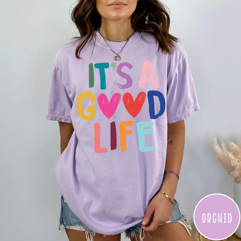 K&ouml;nnte beinhalten: Ein hellviolettes Kurzarm-T-Shirt mit dem mehrfarbigen Schriftzug IT'S A GOOD LIFE, bei dem zwei rote Herzen den Buchstaben O ersetzen. Das Shirt hat einen Rundhalsausschnitt und eine lockere Passform mit einem bunten Grafikdesign auf der Vorderseite.