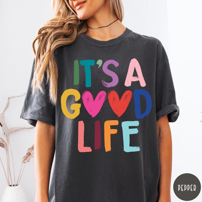 K&ouml;nnte beinhalten: Ein anthrazitfarbenes Kurzarm-T-Shirt mit dem mehrfarbigen Schriftzug IT'S A GOOD LIFE, bei dem zwei Herzformen den Buchstaben O ersetzen. Die Buchstaben sind in verschiedenen Farben gedruckt, darunter Gr&uuml;n, Lila, Pink, Gelb, Rot, Blau und Orange.