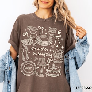 May include: Espresso-coloured t-shirt featuring a white illustration of teacups, plates, and a duck, with the text "I'd rather be thrifting". The shirt has a relaxed, oversized fit. The design is in white.