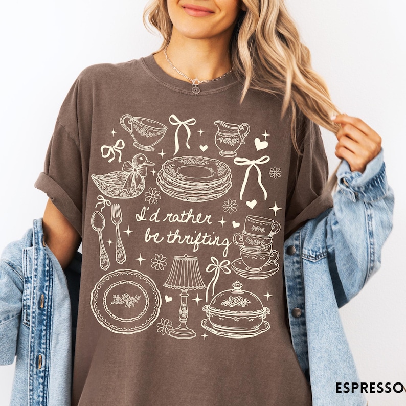 May include: Espresso-coloured t-shirt featuring a white illustration of teacups, plates, and a duck, with the text "I'd rather be thrifting". The shirt has a relaxed, oversized fit. The design is in white.