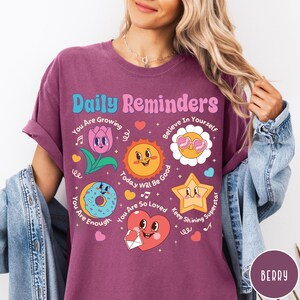 May include: Berry-colored t-shirt with the text "Daily Reminders" and various cartoon graphics. The graphics include a tulip, sun, flower, star, donut, and heart, each with a positive message. The shirt has rolled-up sleeves.