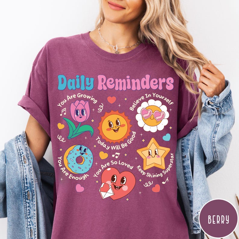 May include: Berry-colored t-shirt with the text "Daily Reminders" and various cartoon graphics. The graphics include a tulip, sun, flower, star, donut, and heart, each with a positive message. The shirt has rolled-up sleeves.