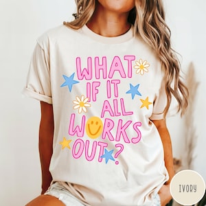 May include: An ivory short-sleeve t-shirt featuring the pink text "WHAT IF IT ALL WORKS OUT?" with a yellow smiley face replacing the letter O. The design includes scattered blue and yellow stars and white daisy flowers across the front.