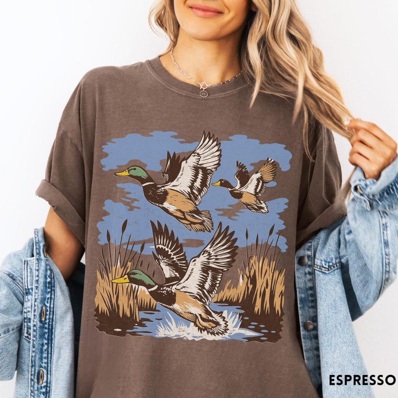 May include: A brown t-shirt with a graphic of three ducks flying over water and reeds. The ducks have green heads, brown bodies, and white wings. The background features a blue sky and brown reeds. The word "ESPRESSO" is printed at the bottom of the shirt.