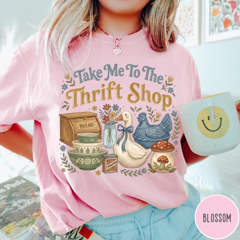 Op de afbeelding: Roze sweatshirt met de tekst "Take Me To The Thrift Shop" en een afbeelding van een eend, kip, brooddoos, kommen en een paddenstoel. Een mok met een smiley wordt in de hand gehouden.
