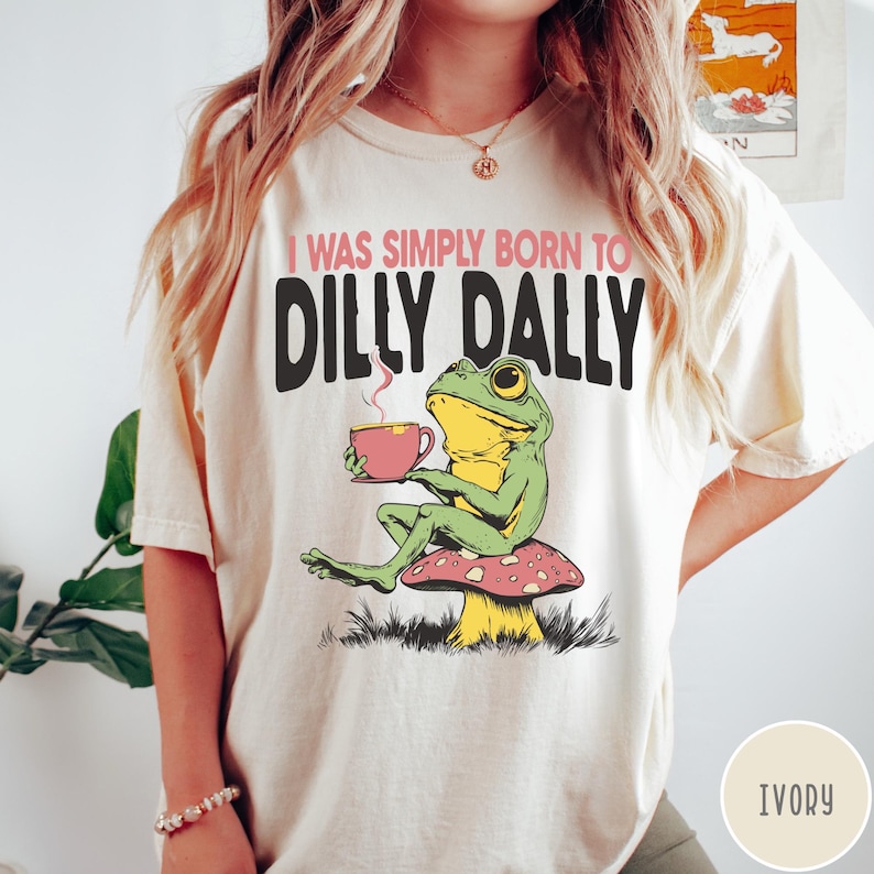 May include: Ivory-colored t-shirt with the text "I WAS SIMPLY BORN TO DILLY DALLY." A cartoon frog is seated on a mushroom, holding a pink teacup. The frog is green and yellow.